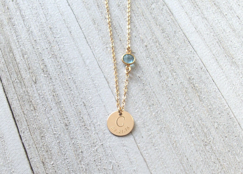 May include: Gold necklace with a round pendant engraved with the letter "C" and the date "08.21.18". A small light blue gemstone is attached to the chain above the pendant.