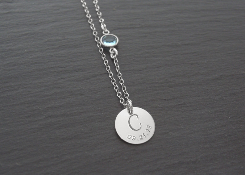 May include: A silver necklace with a round pendant engraved with the letter "C" and the date "09.21.18". A small blue gemstone is attached to the chain above the pendant.