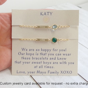 May include: A card with the name "KATY" printed on it. The card has two gold bracelets with small gemstones. The bracelets are labeled "THAI" and "LIEM". The card reads "We are so happy for you! Our hope is that you can wear these bracelets and know that your sweet boys are with you at all times. Love, your Mayo Family XOXO".
