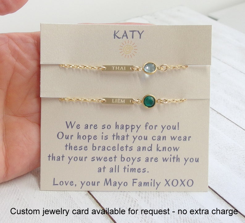 May include: A card with the name "KATY" printed on it. The card has two gold bracelets with small gemstones. The bracelets are labeled "THAI" and "LIEM". The card reads "We are so happy for you! Our hope is that you can wear these bracelets and know that your sweet boys are with you at all times. Love, your Mayo Family XOXO".