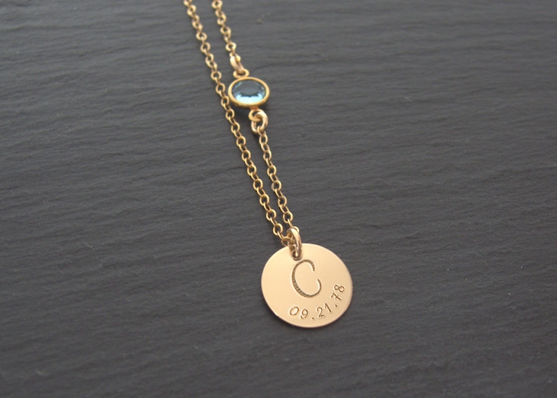 May include: Gold necklace with a round pendant engraved with the letter "C" and the date "09.21.18". A small blue gemstone is attached to the chain above the pendant.