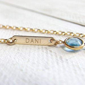 May include: A gold chain necklace with a small blue gemstone charm and a rectangular tag engraved with the name "Dani".
