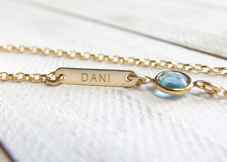 May include: A gold chain necklace with a small blue gemstone charm and a rectangular tag engraved with the name "Dani".