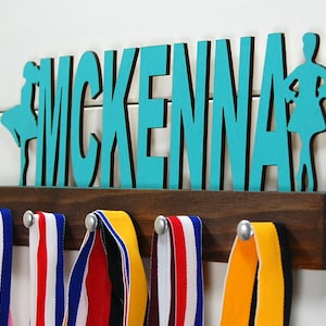 May include: A teal blue wooden medal holder with the name "MCKENNA" and two dancer silhouettes. The holder has six hooks for hanging medals.