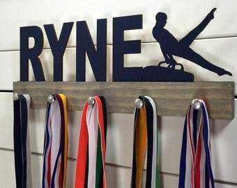 May include: A wooden medal display rack with the name "RYNE" and a silhouette of a gymnast on a pommel horse. The rack has five hooks for hanging medals. There are four colorful ribbons hanging from the hooks.