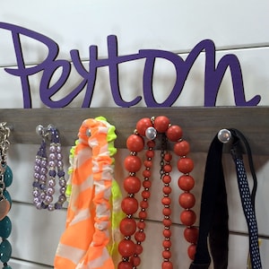 May include: A wooden jewelry organizer with the name "Peyton" in purple lettering. The organizer has hooks for hanging necklaces, bracelets, and other accessories. There are several necklaces, bracelets, and headbands hanging on the organizer.
