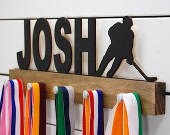Può includere: Un porta medaglie in legno con il nome "JOSH" in lettere nere e una silhouette nera di un giocatore di hockey. Il supporto ha ganci per appendere le medaglie e ci sono diverse medaglie colorate appese ai ganci.