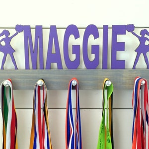 May include: Purple wooden medal holder with the name "MAGGIE" and two cheerleaders on either side. The holder has five hooks for hanging medals. There are four colorful medals hanging on the hooks.