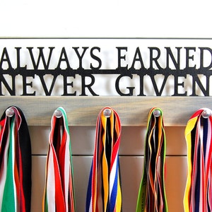 May include: A wooden medal hanger with the words "ALWAYS EARNED. NEVER GIVEN." in black metal lettering. Five colorful medal ribbons are hanging from the hanger.