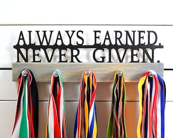 May include: A wooden medal hanger with the words "ALWAYS EARNED. NEVER GIVEN." in black metal lettering. Five colorful medal ribbons are hanging from the hanger.