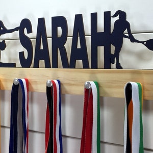 May include: A wooden medal display rack with the name "SARAH" and a silhouette of a tennis player. The rack has five hooks for hanging medals. There are five colorful ribbons hanging from the hooks.