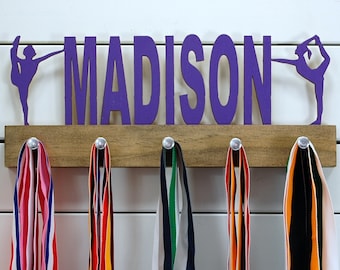May include: A wooden medal hanger with the name "MADISON" in purple letters and two purple gymnast silhouettes. Five medals are hanging from hooks on the hanger.