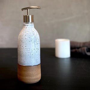 May include: A ceramic soap dispenser featuring a cylindrical body with a two-tone finish. The upper portion is white with black speckles, while the lower base is a solid, unglazed brown clay. It includes a metallic silver pump mechanism on top.