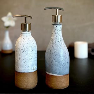 May include: Two ceramic soap dispensers with metallic pumps. The bottles feature a speckled white and gray glaze on the upper portion and a solid, unglazed brown clay base. The dispensers have a cylindrical shape with a rounded shoulder design.