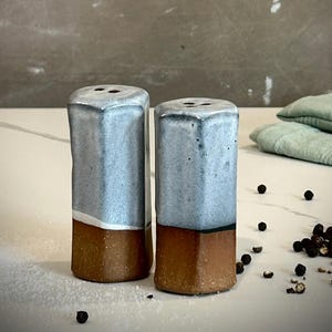 May include: Two cylindrical ceramic shakers featuring a two-tone design. The upper portion is finished in a glossy, speckled light blue glaze, while the lower section remains an unglazed, earthy brown clay. Each top has small dispensing holes.