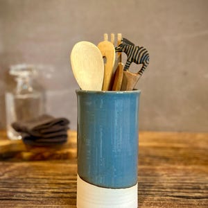 May include: A cylindrical ceramic utensil holder with a glossy blue upper section and a matte white base. It contains several wooden kitchen utensils, including a large spoon, a fork, and a carved wooden server shaped like a zebra with black stripes.