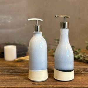 May include: Two ceramic soap dispensers with silver-toned pump mechanisms. Each bottle features a light blue glazed upper section and a solid cream-colored base. The right dispenser includes a thin black horizontal band separating the two colors.