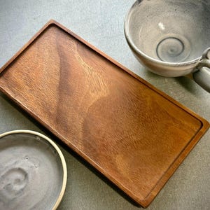 May include: A rectangular wooden serving tray with a smooth, polished finish and visible grain patterns, accompanied by a light gray ceramic mug with a handle and a small, matching light gray ceramic bowl with a smooth, rounded edge.