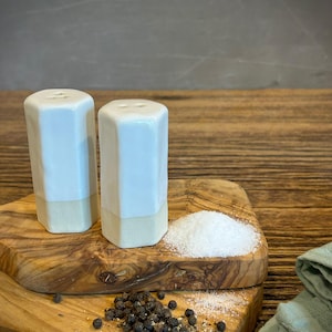 May include: Two hexagonal salt and pepper shakers with a white glazed upper section and a matte, unglazed base. The shakers feature small holes on top for dispensing. A small pile of white salt and scattered black peppercorns sit nearby.