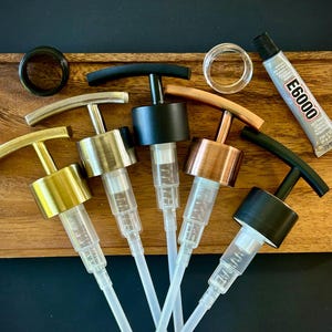 May include: A collection of soap dispenser pumps with various metal finishes, including gold, silver, black, and copper, arranged on a wooden tray. A tube of E6000 glue and additional pump parts are also present.