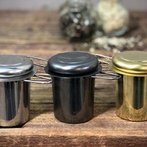 Puede incluir: Tres infusores de t&eacute; con tapas, uno plateado, uno negro y uno dorado, sobre una superficie de madera.