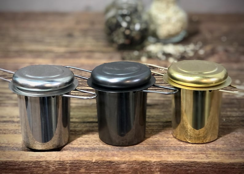 Puede incluir: Tres infusores de t&eacute; con tapas, uno plateado, uno negro y uno dorado, sobre una superficie de madera.