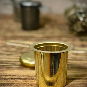 Puede incluir: Un infusor de t&eacute; de metal dorado con un filtro de malla fina. El infusor est&aacute; sobre una superficie de madera.