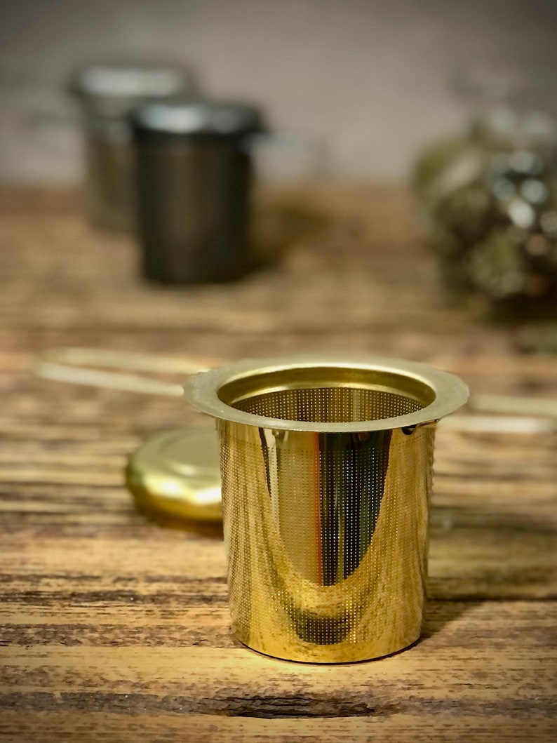 Puede incluir: Un infusor de t&eacute; de metal dorado con un filtro de malla fina. El infusor est&aacute; sobre una superficie de madera.