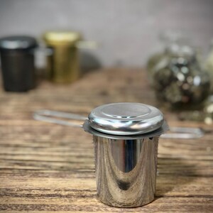 Puede incluir: Un infusor de t&eacute; de acero inoxidable plateado con tapa y asa. El infusor est&aacute; sobre una superficie de madera.