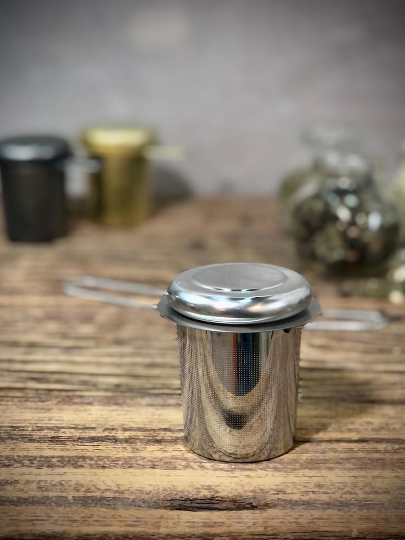 Puede incluir: Un infusor de t&eacute; de acero inoxidable plateado con tapa y asa. El infusor est&aacute; sobre una superficie de madera.