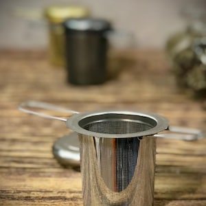 Puede incluir: Un infusor de t&eacute; de acero inoxidable con un filtro de malla fina y un asa. El infusor est&aacute; sobre una superficie de madera.