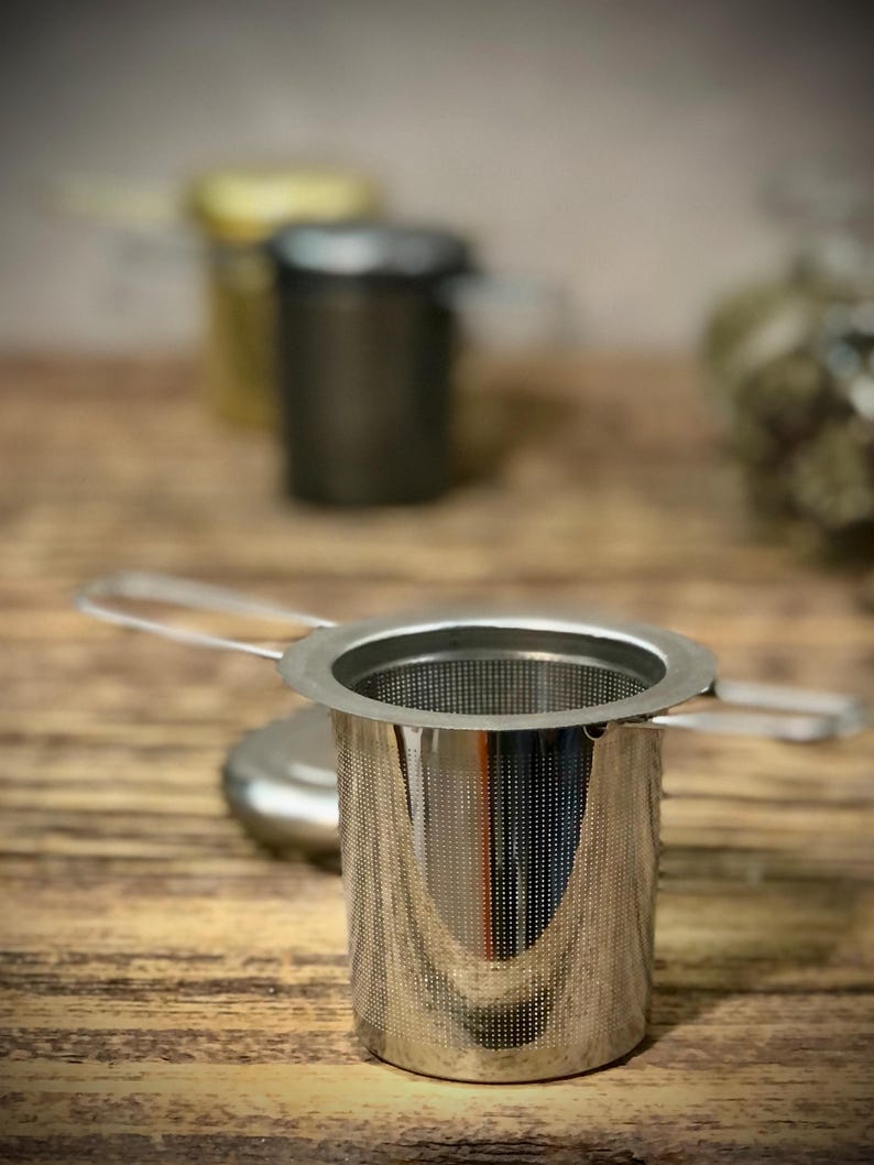 Puede incluir: Un infusor de t&eacute; de acero inoxidable con un filtro de malla fina y un asa. El infusor est&aacute; sobre una superficie de madera.