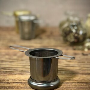 Puede incluir: Un infusor de t&eacute; de plata con un filtro de malla y un asa. El infusor est&aacute; sobre una superficie de madera.
