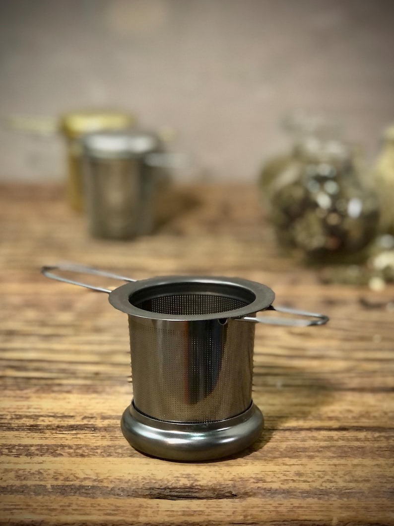 Puede incluir: Un infusor de t&eacute; de plata con un filtro de malla y un asa. El infusor est&aacute; sobre una superficie de madera.