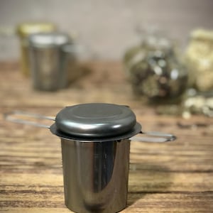 Puede incluir: Un infusor de t&eacute; de acero inoxidable con una tapa negra y un asa. El infusor est&aacute; sobre una superficie de madera.