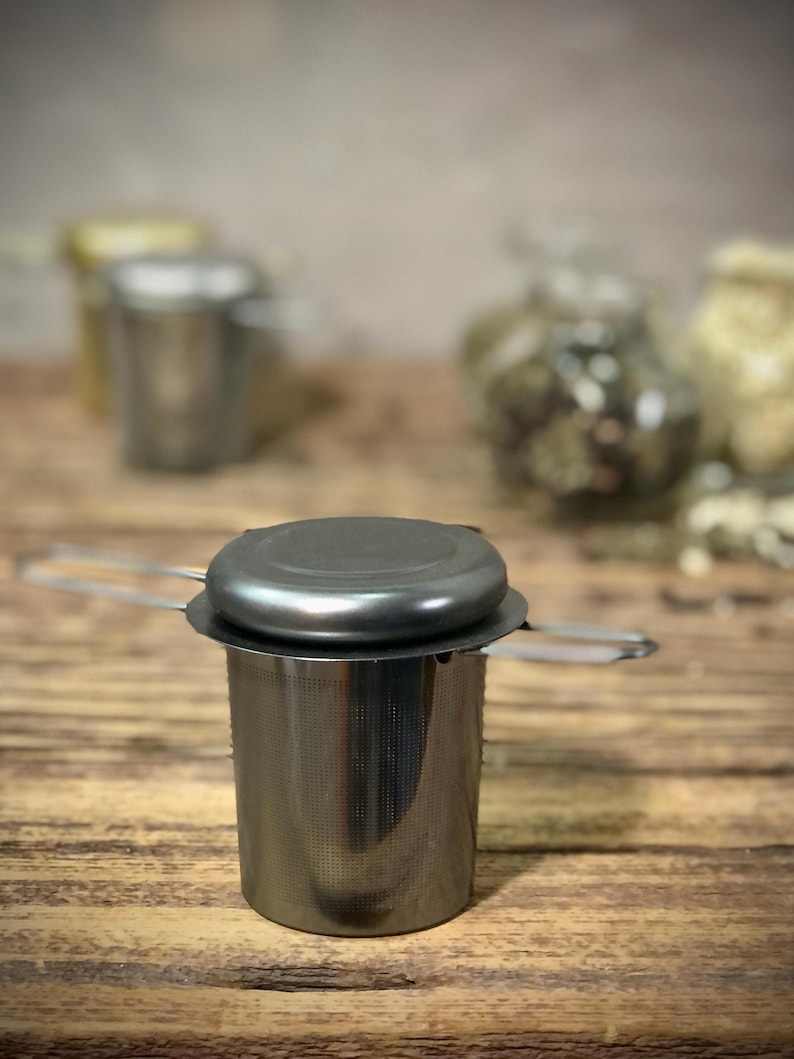 Puede incluir: Un infusor de t&eacute; de acero inoxidable con una tapa negra y un asa. El infusor est&aacute; sobre una superficie de madera.