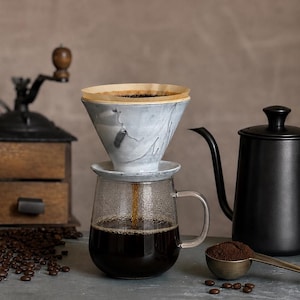 May include: A ceramic coffee dripper with a grey and white marbled pattern sits atop a clear glass mug. A brown paper filter containing coffee grounds is inside the dripper, with dark coffee dripping into the mug. A black gooseneck kettle is nearby.