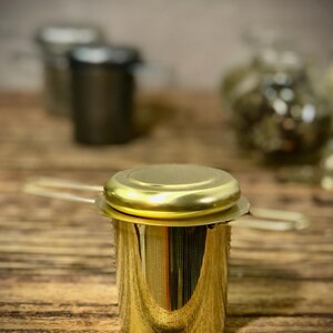 Puede incluir: Infusor de t&eacute; de metal dorado con un filtro de malla y una tapa. El infusor est&aacute; sobre una superficie de madera.