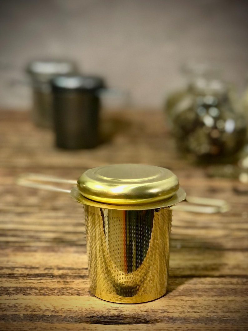 Puede incluir: Infusor de t&eacute; de metal dorado con un filtro de malla y una tapa. El infusor est&aacute; sobre una superficie de madera.