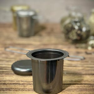 Puede incluir: Un infusor de t&eacute; de plata con un filtro de malla y un asa. El infusor est&aacute; sobre una superficie de madera.