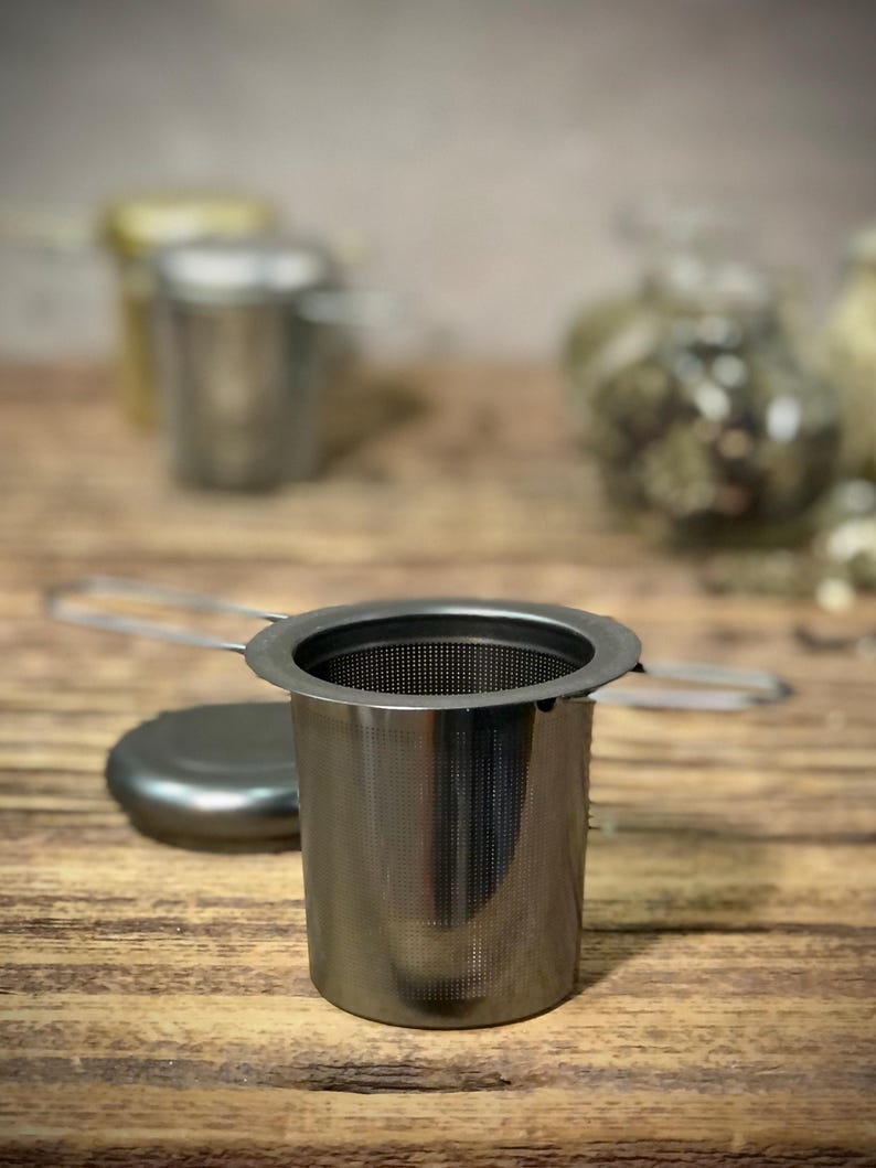 Puede incluir: Un infusor de t&eacute; de plata con un filtro de malla y un asa. El infusor est&aacute; sobre una superficie de madera.