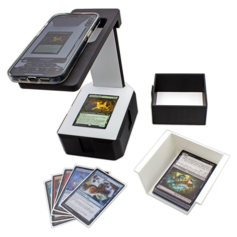 May include: A black and white card scanning device with a smartphone in place. The device has a built-in screen displaying a card image. Several trading cards are fanned out, and a black and white card holder is also present.