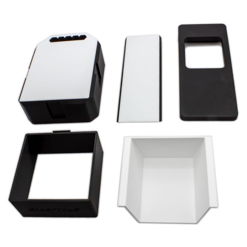May include: An assortment of black and white plastic components, likely for a larger product. The pieces include a rectangular box with a white top, a rectangular frame, and a tray-like structure. The components are arranged against a white background.