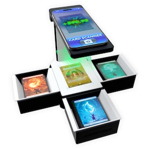 May include: A black and white card scanning station featuring a top-mounted smartphone holder displaying a card scanner interface. The base includes three open compartments holding illustrated trading cards, with a central illuminated scanning area.