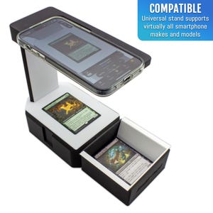 May include: Black and white smartphone stand with a clear phone case holding a phone. The stand is displaying a card game. The text on the stand says "COMPATIBLE" and "Universal stand supports virtually all smartphone makes and models."