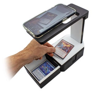 May include: Black and white card scanning device with a smartphone on top. A hand is placing a card on the white platform. The device has a drawer with cards inside. The cards are for a trading card game.