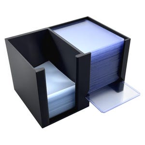 May include: A black plastic storage organizer featuring two distinct compartments. One side holds a stack of clear plastic card sleeves, while the other side contains a larger stack of clear card protectors, with one single protector resting flat.