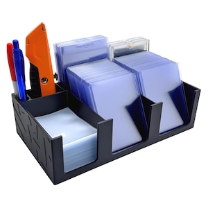 May include: A black plastic desk organizer with a textured geometric pattern on the side. It features multiple compartments holding stacks of clear plastic card sleeves, a blue pen, a red marker, and an orange utility knife with a black grip.