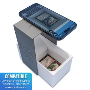 May include: A gray and white phone stand with a phone on top of it. The stand has a slot for cards and the text "Stand" on the side. The phone is displaying a card game app. The text "COMPATIBLE" is on the bottom left corner of the image. The text "Universal stand supports virtually all smartphone makes and models" is below the text "COMPATIBLE".
