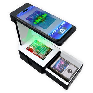 May include: A black and white card scanner stand with a smartphone mounted on top, displaying a digital interface with the text CARD SCANNER. A trading card is positioned on the base below the phone, illuminated by a green light scanning effect.